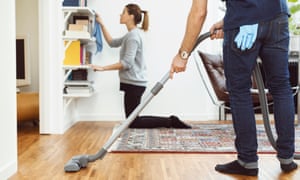 A man vacuums while a woman cleans shelves at home.