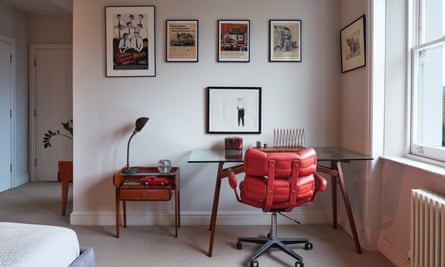The office with a glass-top desk with wooden legs, swivel chair with red padding, and pictures on the wall above