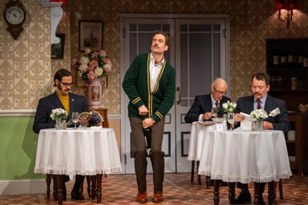 Actors on stage, seated at dining tables in a hotel restaurant, with a man standing centre stage between them.