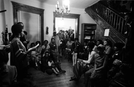 black and white image of a group of people sitting inside a home, looking at one woman speaking