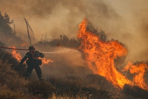 A person in firefighting gears sprays water through a fire hose at a blaze filling the sky with smoke