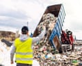 A rear view of a male worker in a hi-vis jacket at a landfill site, directing a dump truck to unload waste