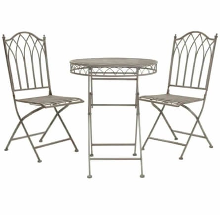 Two grey metal folding chairs. There is an arced pattern in the back of the chairs. In the middle of the chairs is a round folding metal table. The table is grey with a patterned edge.