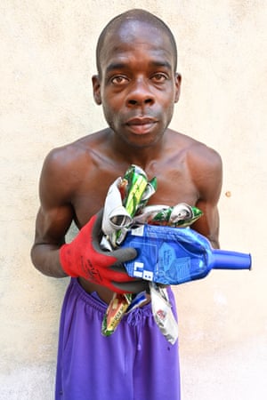 A bare-chested man in purple jogging bottoms holds an armful of crushed cans and bottles