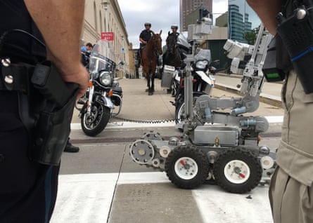 Police officers stand near a remote-controlled robot as horse-mounted officers stand in the background.