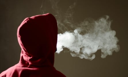 A young male in red hoodie facing away form the camera, vaping and exhaling a large cloud of white vapour
