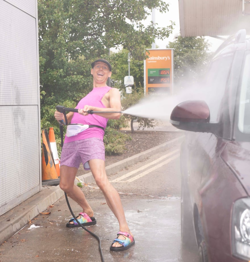 Tim Jonze using a jet-washer on his car in a Sainsbury’s garage, while wearing a bright pink vest, shorts and sandals