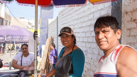 A woman wearing an apron with a man next to her stands under a parasol smiling at the camera with a metal bowl of fish fillets next to her while a customer looks on.
