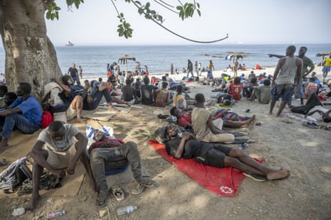 People sit or lie on the ground near a tree on a beach