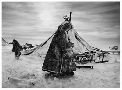 A woman and child dressed in animal skins stand on thick snow in front of a tent.