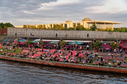 People sitting in red deckchairs lined up by the Spree River