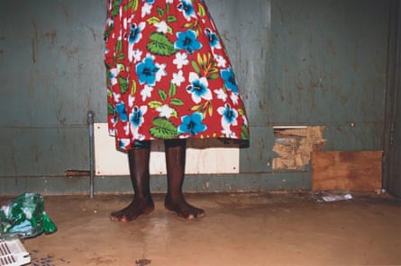 Une vue de Nancy Kidd de la taille vers le bas alors qu'elle se tient devant un mur endommagé dans sa maison.  Elle est pieds nus et porte une jupe rouge vif avec des fleurs bleues et blanches dessus