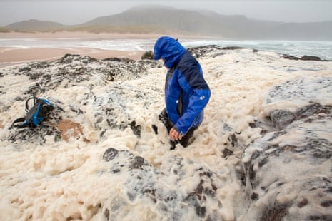 A figure in blue waterproof jacket knee-deep in sea foam during gale force winds on the beach.