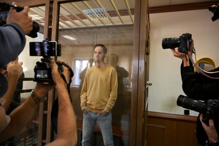 a man in a sweatshirt and jeans stands behind glass as photographers take pictures