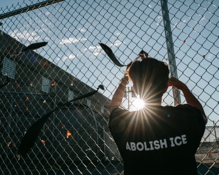 A protester ties a ribbon on a mesh fence surrounding Delaney Hall, wearing a shirt that says 'Abolish ICE'