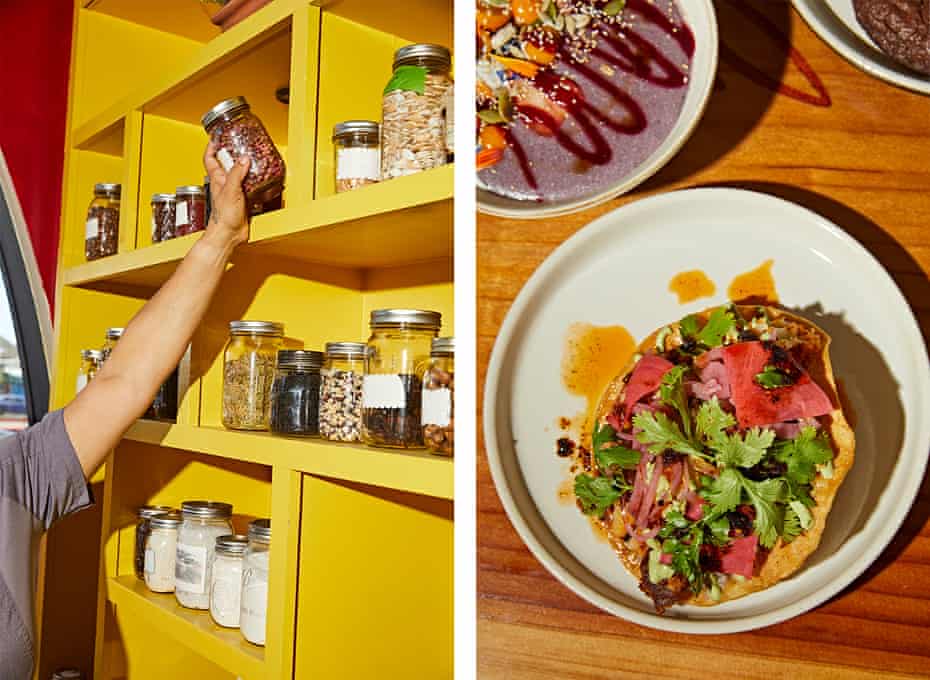 Bright yellow shelves hold jars of grains and seeds as a woman’s hand reaches up to select a jar.