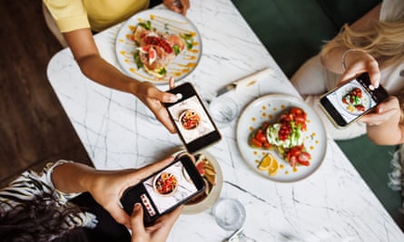 From a top-down perspective, friends gather around a marble table, using their smartphones to photograph colorful and beautifully presented dishes at a chic cafe.