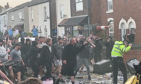 Protesters shout and throw objects at a police officer in a residential street with broken debris on the ground.