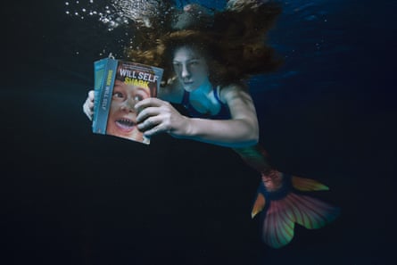 The author reading a book underwater while wearing an orange mermaid tail.