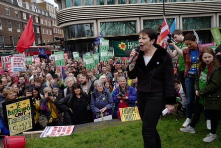 Lucas speaks animatedly into a microphone before a crowd of demonstrators holding placards and flags.
