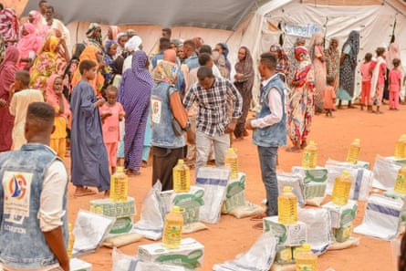 Sudanese people milling about near small piles of cooking oil, pasta and other food in an open area near a tent with a Unicef banner