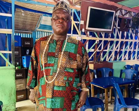 ‘They told us to leave. They didn’t tell us where to go’: the demolitions destroying homes and lives in Lagos A Nigerian man wearing a red, green and gold patterned top and traditional hat standing in a room next to blue plastic chairs and tables