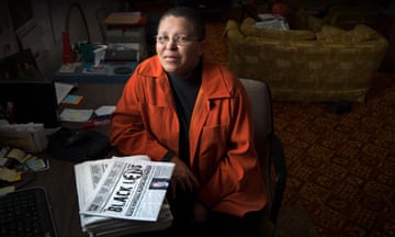 Person wearing orange jacket sits at desk in front of stack of newspapers
