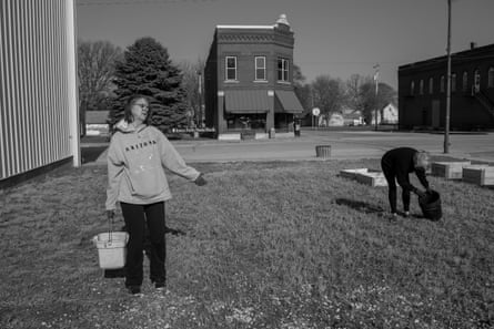 Two women with buckets work in a square of grass off a main street in a rural town. Behind them are several raised garden beds.