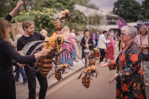 Two people are manipulating giant bee puppets, with a woman in a floral dress in front of them