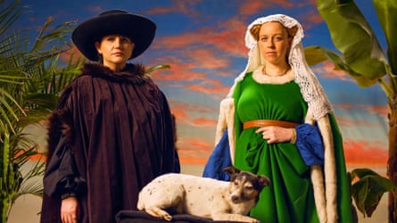 Natasia Demetriou and Ellie White in historical costume stand with a dog against a painted sky backdrop.