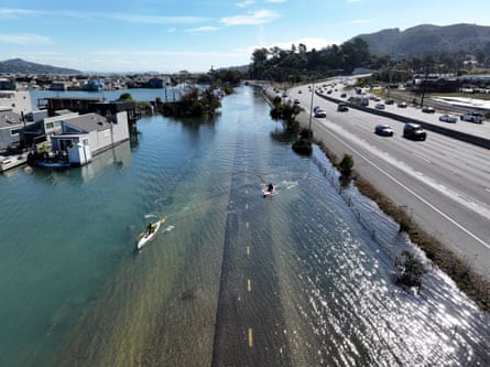 Kayakers paddle a flooded road beside houseboats, next to a busy Marin County highway