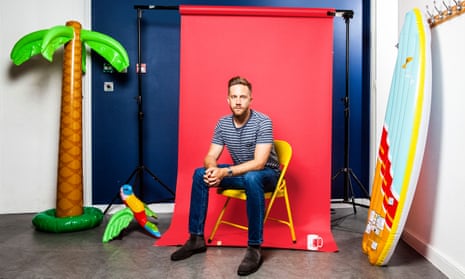 Tom Blomfield, in jeans and a t-shirt, sitting on a yellow metal chair, a red screen behind him, and with a blow-up palm tree and parrot one side of him, and a blow-up surf board propped up the other