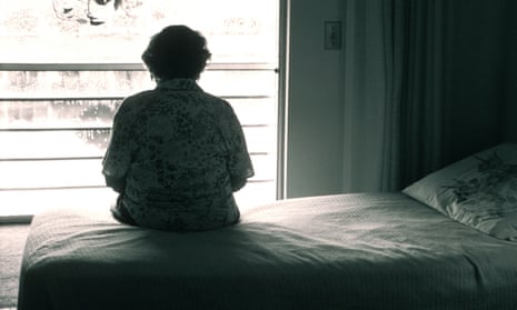 An elderly woman sitting on her bed looking out a window
