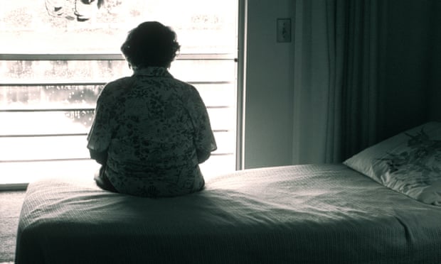 An older woman sits on a bed looking out a window