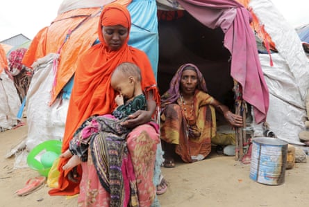 A woman in an orange shawl and headscarf cradles a child outside a tent where an older woman crouches by the entrance
