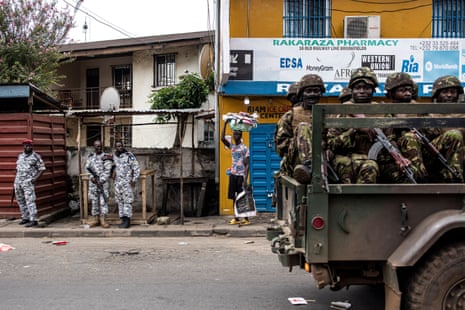 A soldiers in camouflage gear on the back of a vehicle, carrying assault rifles, pass soldiers on the street