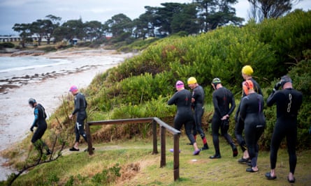 Group of ocean swimmers in wet suits and swimming caps walking down a hill and towards the beach shore.