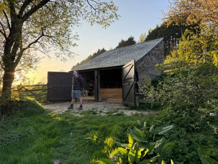 A man standing by a tree outside a bothy with big open double doors at sunset