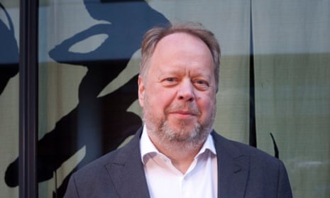 Portrait of Andy Palmer in a suit and open-necked shirt standing by a window with an abstract black design on the glass