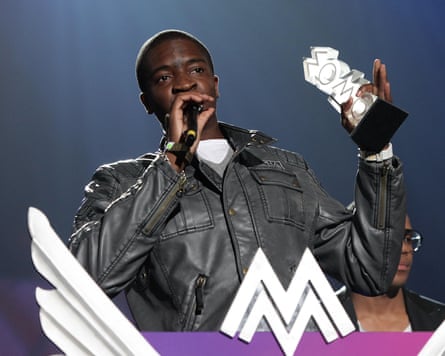 Guvna B speaks into a microphone on stage holding his Mobo awards.
