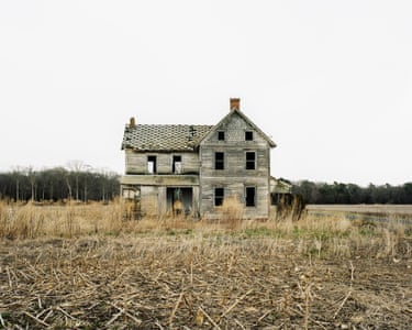 a large clapboard farmhouse, three levels tall with a verandah, covered porch, and a shingled top storey. Broken windows allow you to see right through upstairs