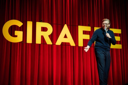 Rob Beckett in dark top and trousers doing his standup routine against a red curtain with big title in yellow capital letters reading Giraffe