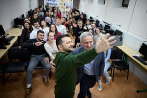 Volodymyr Zelenskiy holds up a smartphone to take a picture of students sitting behind. A man in a suit is stood next to Zelenskiy with his arm round him