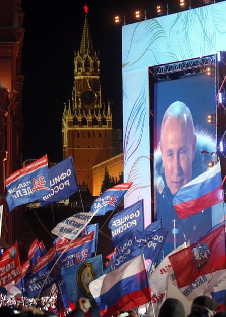 People hold flags in front of a screen showing Vladimir Putin with the Kremlin in the background
