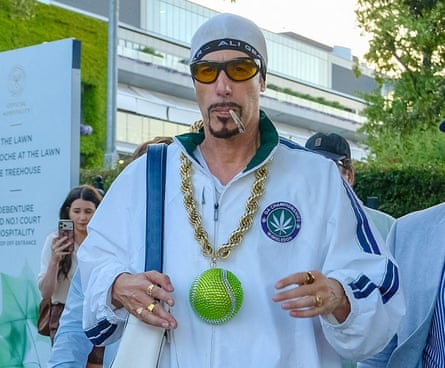 Sacha Baron Cohen in character as Ali G, wearing, sunglasses and a white tracksuit top with Wimbledon logo and a giant green tennis ball on a chunky gold chain round his neck