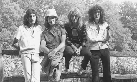 A black and white image of Freddie Mercury, John Deacon, Roger Taylor and Brian May either standing against or sitting on a wooden fence.