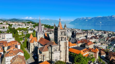 Aerial view of a cathedral in a town with Gothic towers and terracotta rooftops with a view of Lake Geneva and the Alps