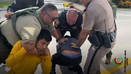 Four officers apprehend two men. One officer holds a man on his knees in a chokehold, while he and two of the other officers push downward on a man crouching on the ground.