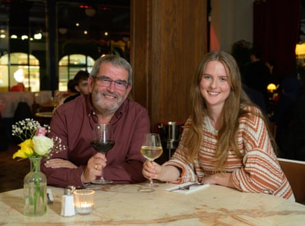 Steve and Eva sitting at a restaurant table holding glasses of wine and smiling at the camera