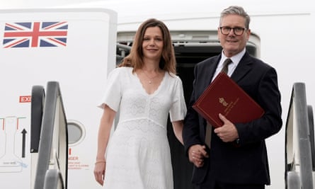 A woman in a white dress and a man in a suit stand holding hands when leaving an aeroplane